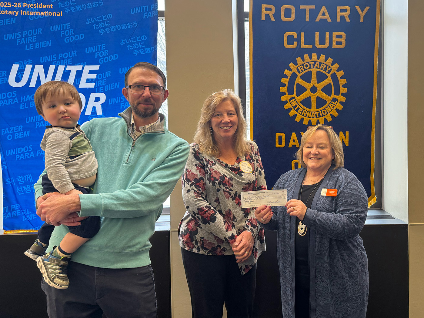 A man holding a baby stands with two women holding a check in front of Rotary flags.